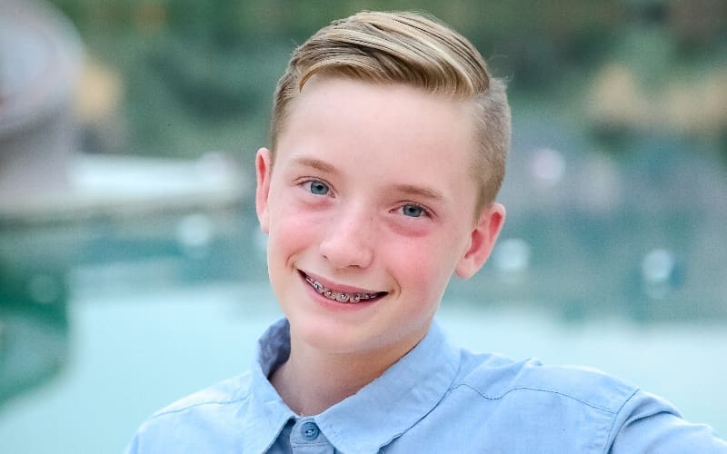 A smiling boy with braces and short, neatly combed hair is wearing a light blue button-up shirt. The background is blurry, showcasing a calm outdoor setting.