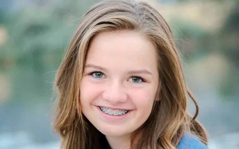 A young girl with long brown hair smiles, showing her braces. The background is blurred, emphasizing her happy expression. She wears a blue top.
