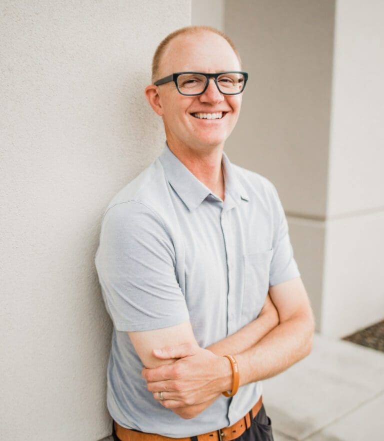 A smiling man in glasses stands against a white wall with arms crossed. He wears a light gray polo shirt and dark pants. The background shows part of a concrete area and building corner.