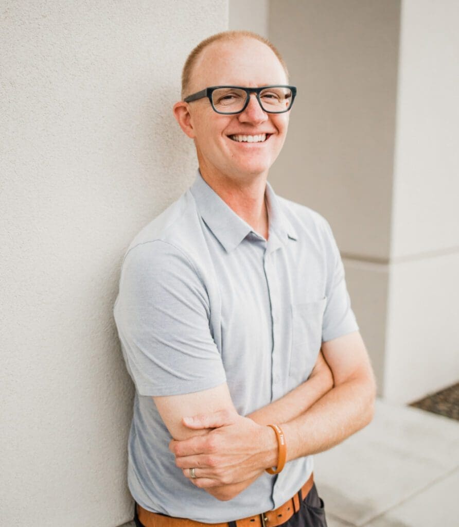 A smiling man in glasses stands against a white wall with arms crossed. He wears a light gray polo shirt and dark pants. The background shows part of a concrete area and building corner.