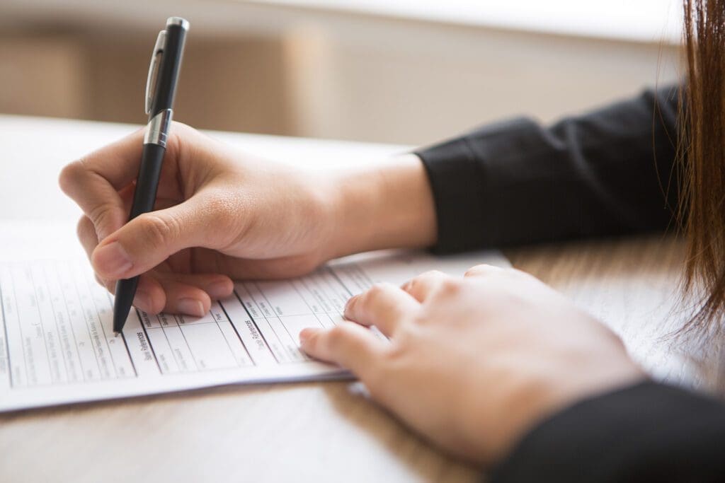 A person with long hair is filling out a form on a wooden table, holding a pen with their right hand. The focus is on the hand and the form, which has several fields and lines. The person is wearing a black sleeve.