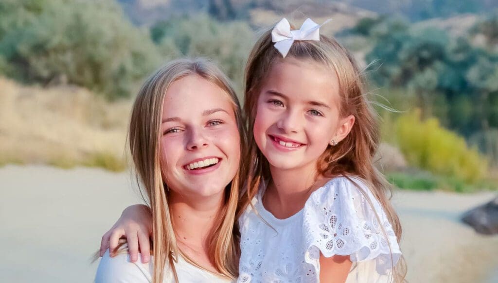 A girl and a young woman with long blonde hair smile brightly. The girl wears a white dress with a bow in her hair, while the woman is in a white top. They are outdoors with blurred greenery in the background.