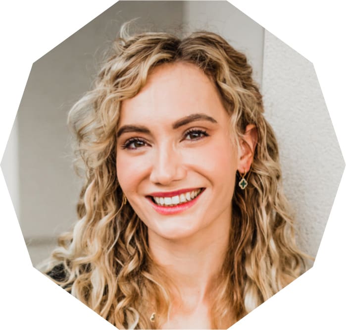 A person with long, wavy blonde hair smiles warmly at the camera. They are wearing earrings and a light-colored top. The background is neutral, focusing attention on their face.