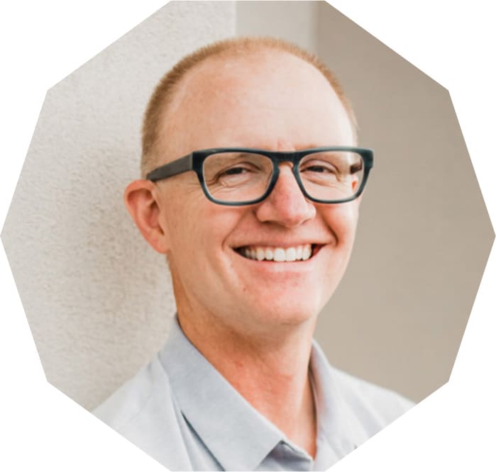 A man with glasses and a light shirt is smiling while standing against a light-colored wall. The image is cropped into an octagonal shape.