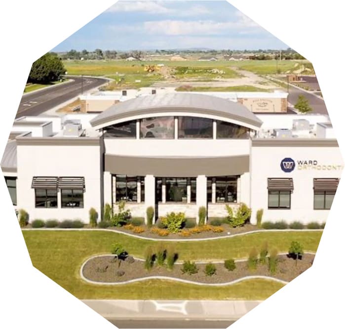 A modern office building with a curved roof under a partly cloudy sky. The building is surrounded by landscaped greenery and situated near a road, with open fields visible in the background.