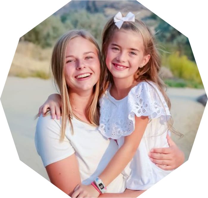 Two smiling girls pose together outdoors. The older girl holds the younger, who wears a white dress with a bow in her hair. They both appear happy, set against a blurred natural background.
