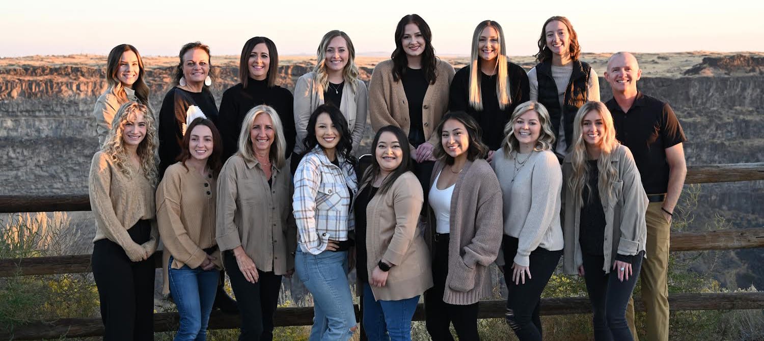 A group of sixteen people standing outdoors in front of a wooden fence and a scenic canyon backdrop. They are all smiling and dressed in casual attire, enjoying the natural setting at sunset.