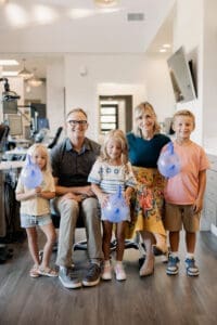 A smiling family of five in an office, with children holding blue medical gloves inflated like balloons, illustrating the benefits of early orthodontic treatment. The children are with Dr Ward and Dr Campbell in Twin Falls, ID.